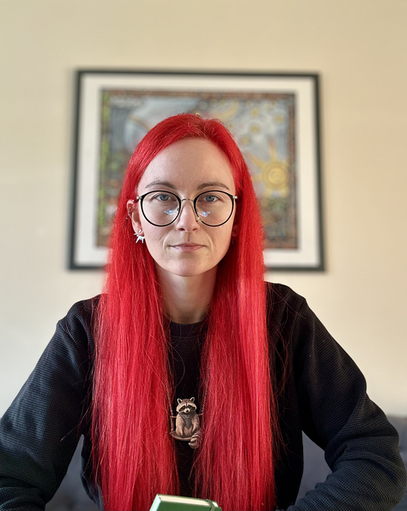 White person with long, red hair, wearing classes and looking at the camera. They are wearing a black top, holding a green notebook, and wearing a racoon pendant.